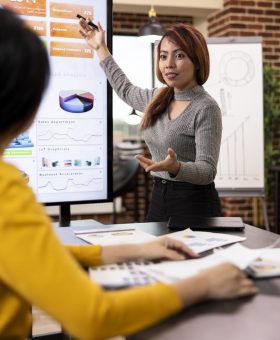 Marketing consultant standing and presenting growth data to coworker seated at office desk. Female business advisor gesturing to device screen, showing her colleague the company analytics summary.
